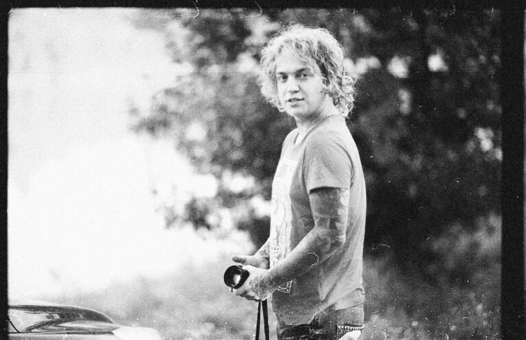 Black and white portrait of a tattooed photographer outdoors holding a camera.